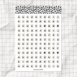 以下が含まれることがあります： タイプライターのキーデザインのアルファベット文字ステッカーシート。白いステッカーには、ハートや星を含む黒い文字と記号があります。シートはグリッドパターンの背景で、「dessinly studio」というテキストがあります。