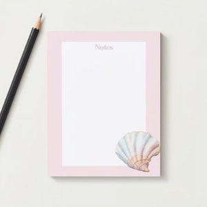 以下が含まれることがあります： ピンクのメモ帳に白い貝殻のイラストと「Notes」という文字が上部に印刷されています