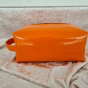 Könnte beinhalten: Eine leuchtend orangefarbene, glänzende, rechteckige Kosmetiktasche mit einem schwarzen Reißverschluss und einem schwarzen Griff.