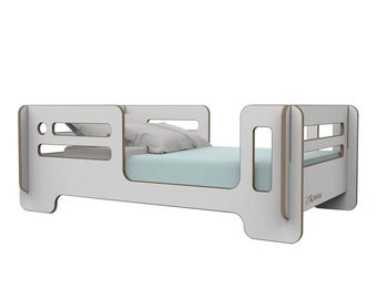 Può includere: Letto per bambini bianco con sponde di sicurezza e un materasso con motivo blu chiaro. Il letto ha un design a taglio sulla testata e sul piede. Il nome del marchio "Trans" è visibile sul piede.
