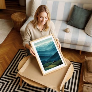 May include: A framed art print featuring a landscape of layered blue and green mountains. The artwork is in a light wood frame and is being held by a person. The print is on a round table with a black and white rug.