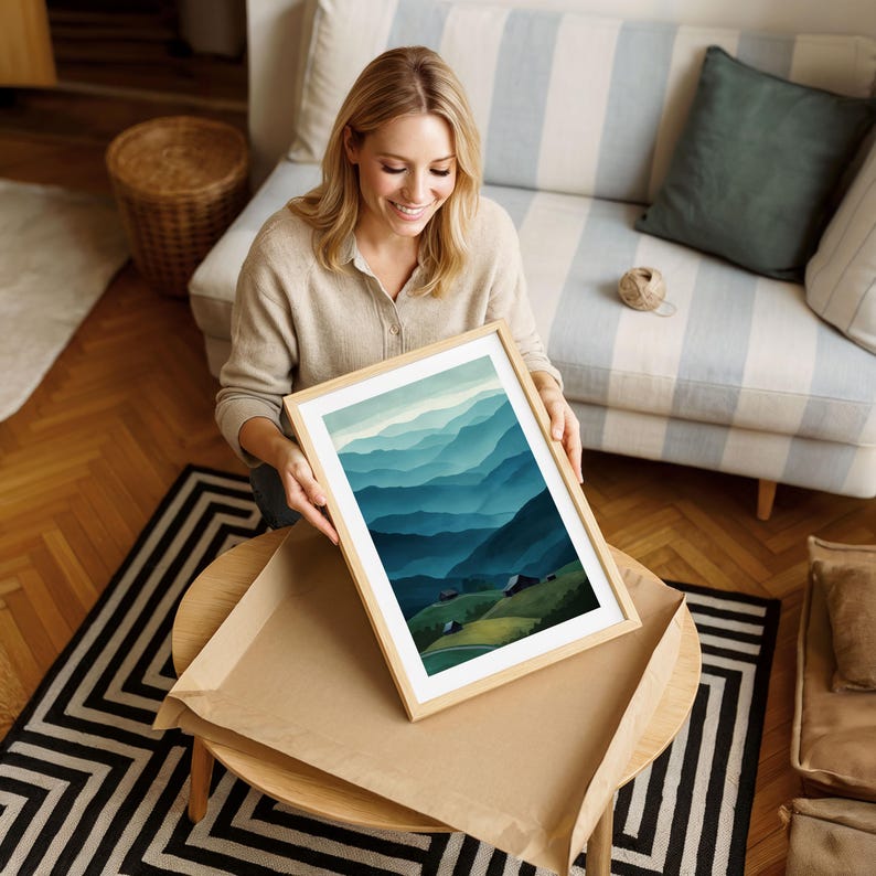 May include: A framed art print featuring a landscape of layered blue and green mountains. The artwork is in a light wood frame and is being held by a person. The print is on a round table with a black and white rug.