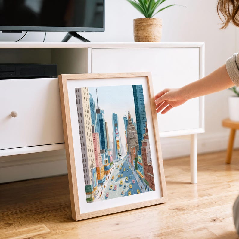 K&ouml;nnte beinhalten: Ein gerahmter Kunstdruck, der eine lebhafte Stadtlandschaft mit hohen Geb&auml;uden, Werbetafeln und gelben Taxis zeigt. Das Kunstwerk befindet sich in einem hellen Holzrahmen und lehnt an einem wei&szlig;en Schrank. Die Farbpalette umfasst Blau-, Rosa- und Gelbt&ouml;ne.