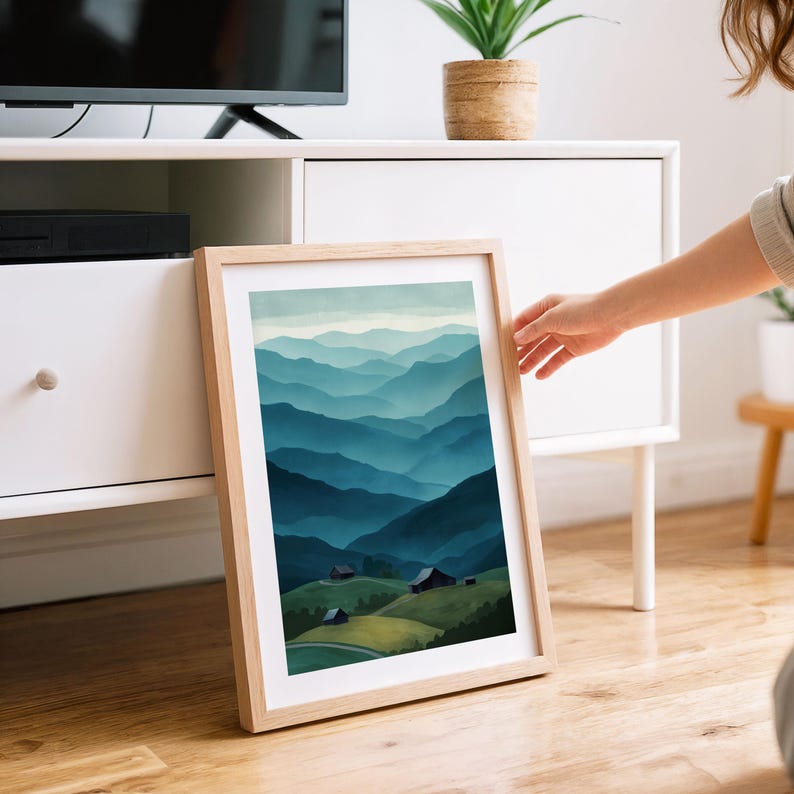 May include: A framed art print featuring a watercolor landscape of layered blue mountains. The artwork includes small buildings on green hills in the foreground. The frame is made of light-colored wood.