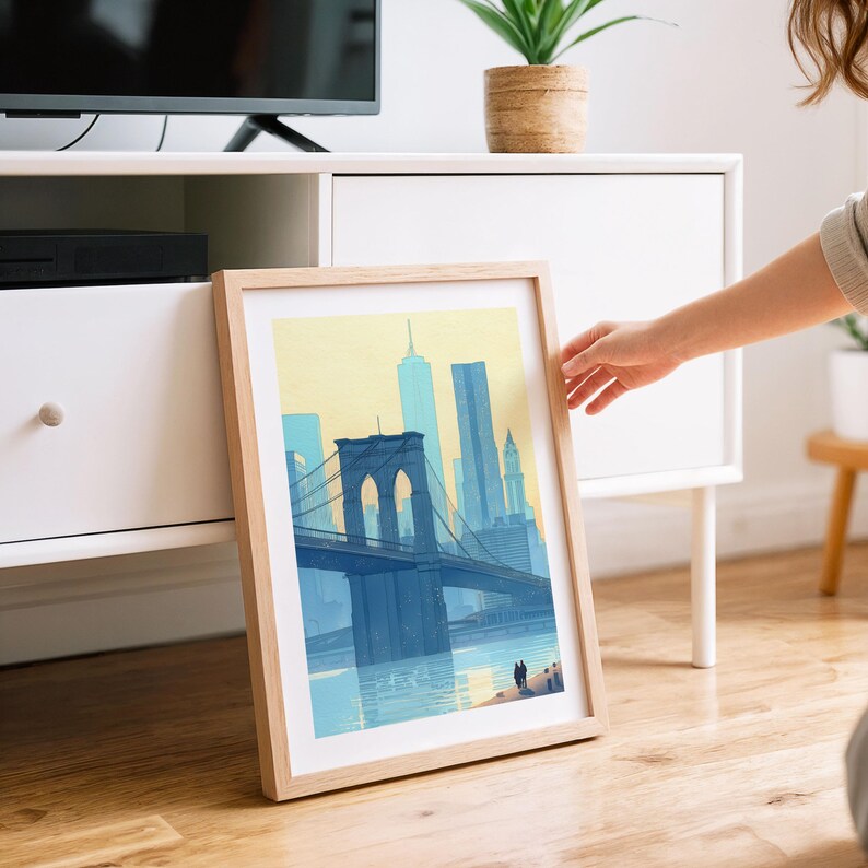 Puede incluir: Una impresi&oacute;n art&iacute;stica enmarcada que presenta un paisaje urbano azul y amarillo con el puente de Brooklyn. La obra de arte tiene un marco de madera clara y representa el puente, los edificios y el agua. La impresi&oacute;n est&aacute; apoyada contra un mueble blanco sobre un suelo de madera.