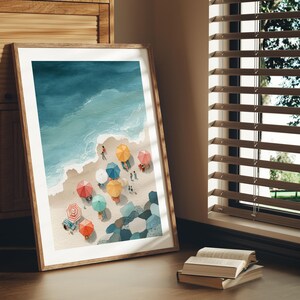 May include: A framed art print depicts a beach scene with colorful umbrellas and people. The artwork features a top-down view of the ocean, sandy shore, and beachgoers. The umbrellas are in various colors, including red, yellow, and blue. The frame is made of wood.
