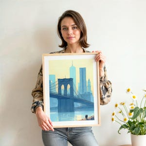 Puede incluir: Una impresi&oacute;n art&iacute;stica enmarcada con una ilustraci&oacute;n azul y amarilla del puente de Brooklyn y el horizonte de la ciudad de Nueva York. La obra de arte est&aacute; sostenida por una persona. El marco es de madera clara.