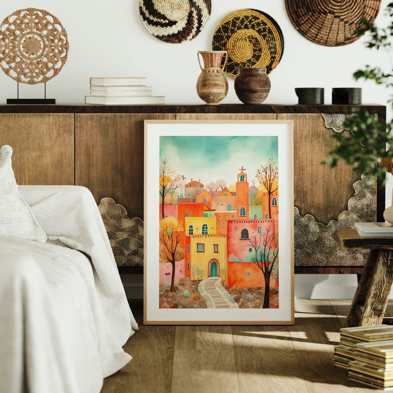 May include: A watercolor painting of a colorful village with a church steeple, surrounded by trees and a winding path. The painting is framed in a light wood frame and is hanging on a wall.