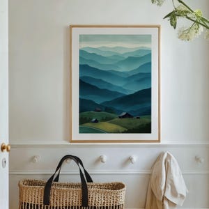 May include: A framed watercolor print depicts a mountain landscape in shades of blue and green. The artwork features layers of rolling hills and distant mountains, with small buildings nestled in the valleys. The frame is a light wood color.