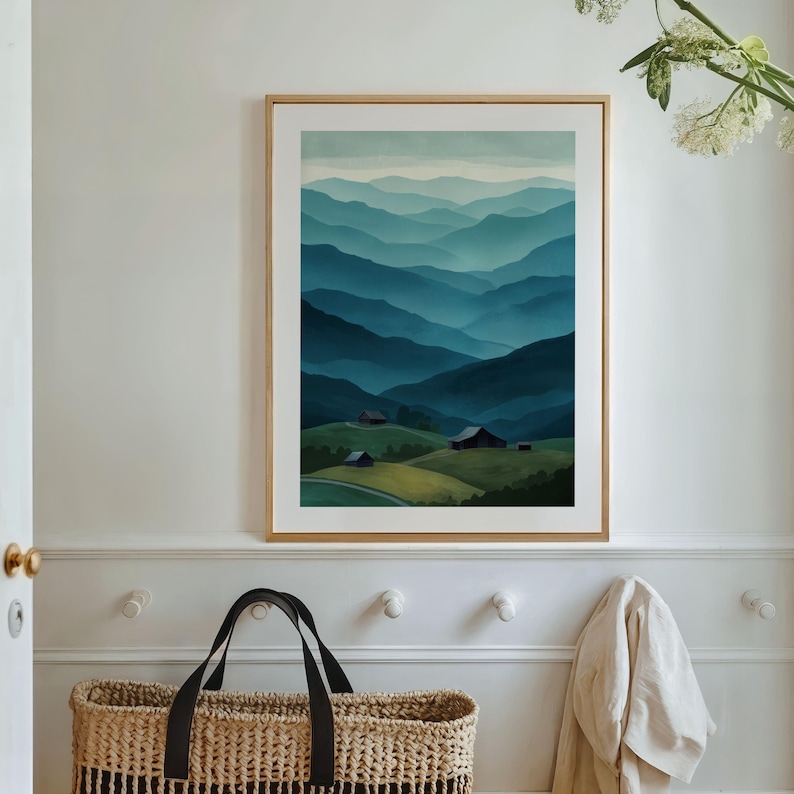 May include: A framed watercolor print depicts a mountain landscape in shades of blue and green. The artwork features layers of rolling hills and distant mountains, with small buildings nestled in the valleys. The frame is a light wood color.
