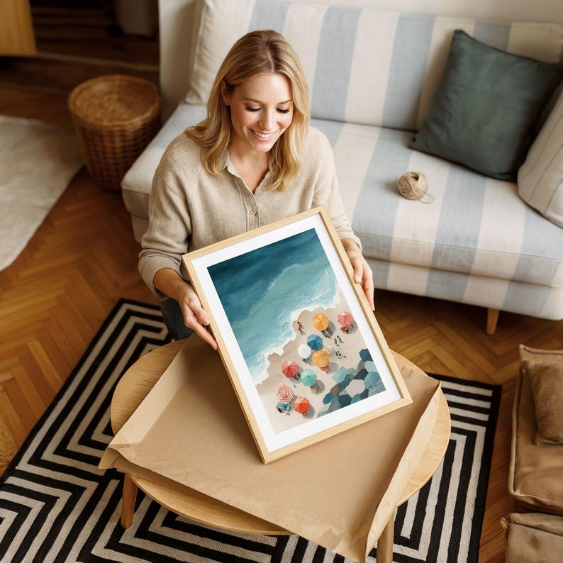 May include: A framed art print depicting a beach scene with colorful umbrellas, ocean waves, and a sandy shore. The artwork is in a light wooden frame. The image is being held by a person.