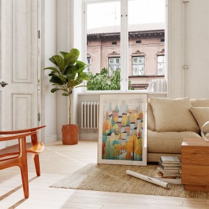 May include: A framed illustration of a colorful cityscape with buildings and trees in shades of green, yellow, orange, and pink. The illustration is hanging on a white wall in a living room setting with a couch, a plant, and a rug.