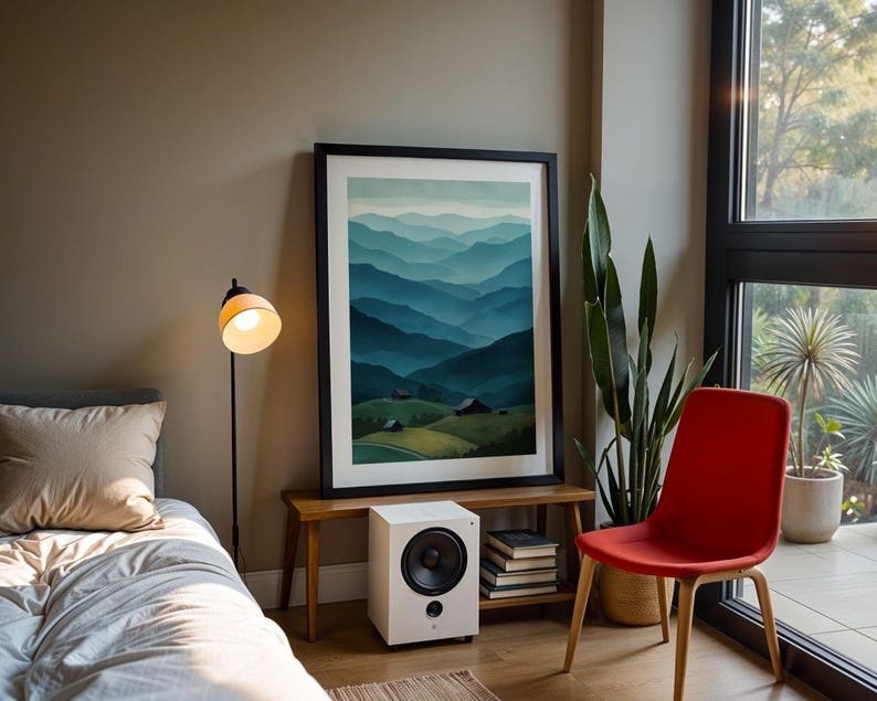 May include: A framed landscape art print depicts layered blue mountains under a light blue sky. A white speaker, books, and a floor lamp with a yellow lampshade are also visible. A red chair and a large plant are near a window.