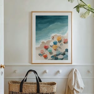 May include: A framed watercolor print depicts a beach scene with colorful umbrellas, people, and the ocean. The artwork features a sandy shore, turquoise water, and a variety of brightly colored beach umbrellas, creating a vibrant and inviting coastal theme.