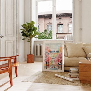 May include: A framed watercolor painting of a city street scene with pink and white flowering trees. The painting is in a gold frame and is hanging on a white wall.