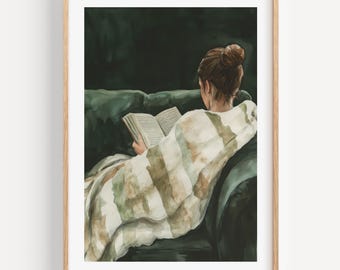 May include: Watercolor print of a person reading a book while sitting on a green sofa. The person is wrapped in a striped blanket with shades of green and brown. The artwork is framed in a light wood frame.