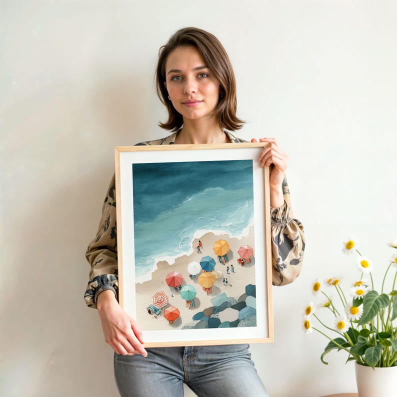 May include: A framed art print depicting a beach scene with colorful umbrellas, people, and the ocean. The artwork features a light wooden frame and a white mat. The print showcases a vibrant beach with blue water and a sandy shore.