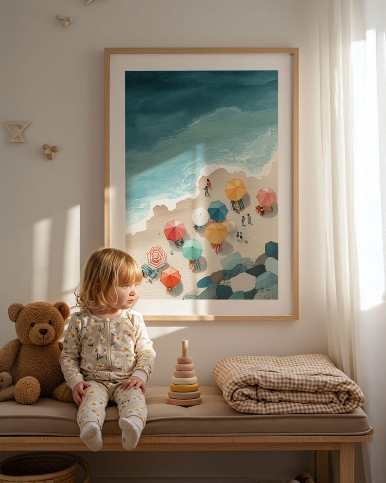 May include: A framed watercolor print depicts a beach scene with colorful umbrellas and people. A young child in a daisy-print onesie sits on a bench next to a teddy bear, a wooden stacking toy, and a folded blanket. The artwork is in a light wood frame.