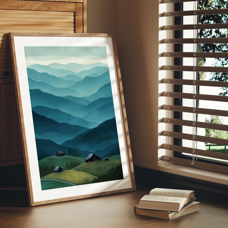 May include: A framed art print featuring a watercolor landscape of layered blue mountains. The artwork depicts rolling hills with small buildings in the foreground. The frame is made of light-colored wood, and the print is leaning against a wall near a window.