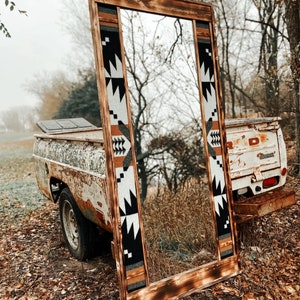 Peut inclure: Un grand miroir en bois avec une finition rustique brûlée. Le miroir est encadré d'un tissu à motifs géométriques noir et blanc. Le miroir est appuyé contre une benne de camion rouillée.