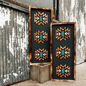 Könnte beinhalten: Zwei Holzschildchen mit südwestlichem Design. Die Schilder zeigen ein geometrisches Muster mit türkisfarbenen und braunen Akzenten. Die Schilder hängen an einer rustikalen Wand.