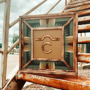Può includere: Un cartello in legno incorniciato con un motivo geometrico in tonalità di verde, marrone e beige. Il cartello presenta una lettera stilizzata "C" con una linea curva sopra, il tutto in un colore arancione bruciato.