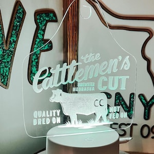 May include: A white acrylic sign shaped like a cattle ear tag with the text "The Cattlemen's Cut Brewster Nebraska" and a silhouette of a cow. The sign has the text "Quality Bred On" and "Not Fed On" on the bottom.