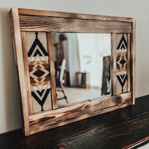 Puede incluir: Un espejo de madera con un acabado quemado y una incrustación de tela con un patrón geométrico. El espejo es rectangular y tiene un marco simple.