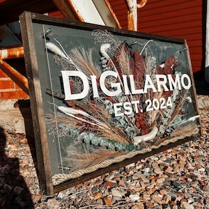 May include: A wooden framed sign with a dark gray background and a white floral arrangement. The sign says "DIGILARMO EST. 2024".