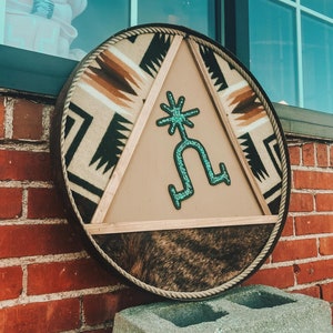 May include: A round, wooden sign with a brown, white, and black patterned border. The center of the sign features a turquoise blue stylized design of a horseshoe with a star on top. The sign is mounted on a brick wall.