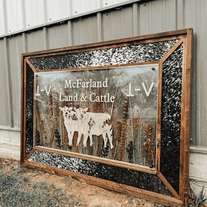 May include: A wooden framed sign with a black and white cowhide background. The sign has a clear acrylic insert with a silhouette of three cows and the text "McFarland Land & Cattle L-V".