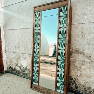Puede incluir: Un espejo con marco de madera con un patrón geométrico en tonos de verde, blanco y rojo. El espejo refleja un cielo azul y un edificio en la distancia.