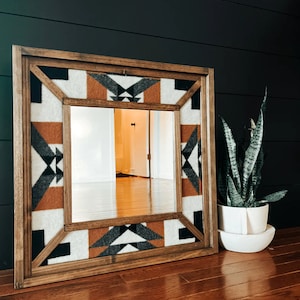 May include: A wooden framed mirror with a geometric pattern in shades of brown, white, and gray. The mirror is reflecting a room with hardwood floors.