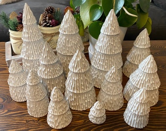 May include: A collection of textured, white ceramic Christmas trees of varying sizes arranged on a wooden table. The trees have a layered design, resembling a carved appearance. The scene suggests a festive, holiday theme.