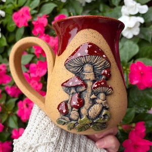 May include: A ceramic mug with a speckled tan base and a deep red glazed rim. The front features a raised, three-dimensional relief of several mushrooms with red caps and white spots, set against a textured tan background with green foliage.