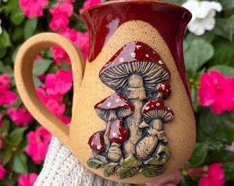 May include: A ceramic mug with a speckled tan base and a deep red glazed rim. The front features a raised, three-dimensional relief of several mushrooms with red caps and white spots, set against a textured tan background with green foliage.