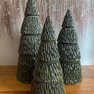 May include: Three decorative ceramic Christmas trees in varying sizes. The trees are dark green with a textured, layered design, and sit on a wooden surface. The background is a neutral color.