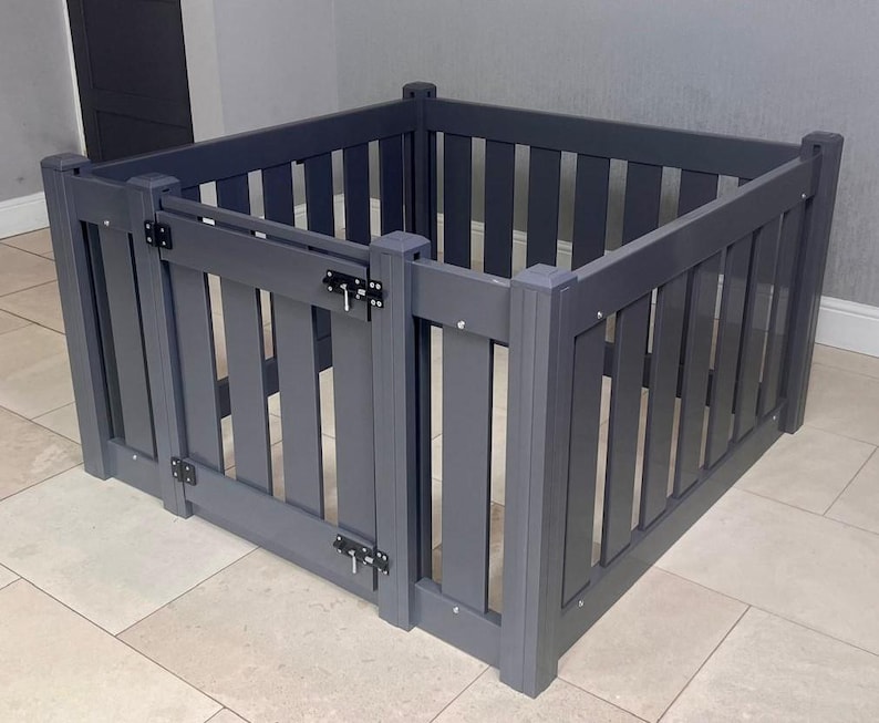 Może przedstawiać: Szary drewniany kojec dla zwierząt z pionowymi listwami i drzwiami na zawiasach. Kojec ma kształt kwadratu i posiada metalowe zatrzaski i zawiasy dla bezpiecznego zamknięcia. Prosty i funkcjonalny design.