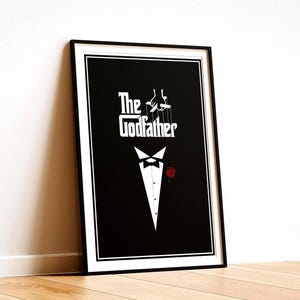 以下が含まれることがあります： 黒い背景に白い文字で「The Godfather」と書かれた額縁入りの長方形のポスター。テキストの上には白いマリオネットの手のグラフィックがあり、その下には黒い蝶ネクタイと小さな赤いバラが描かれた白いタキシードシャツのフロント部分がデザインされています。