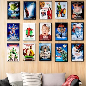 May include: A collection of framed movie posters featuring Christmas-themed films. Posters include "The Grinch," "Elf," "Home Alone 2," and "The Santa Clause." The posters are displayed on a light wood-paneled wall.