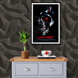 May include: A black framed movie poster with a skull graphic and the title "LAID TO REST" in red text. The poster is hanging on a wall with a dark gray textured wallpaper. A small potted plant and a red and white bowl are on a gray dresser with two drawers.