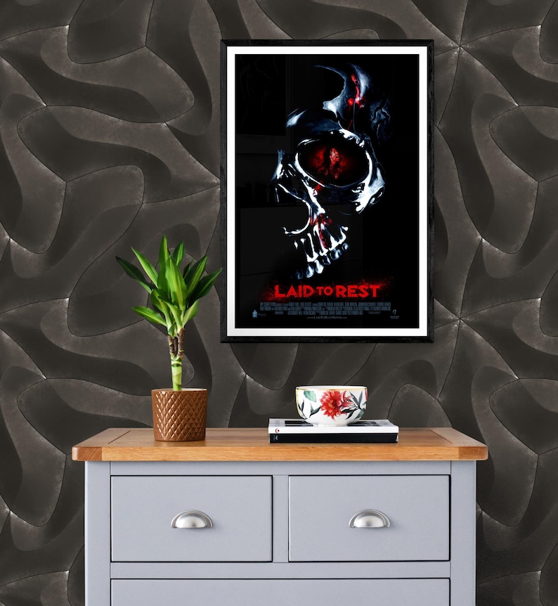 May include: A black framed movie poster with a skull graphic and the title "LAID TO REST" in red text. The poster is hanging on a wall with a dark gray textured wallpaper. A small potted plant and a red and white bowl are on a gray dresser with two drawers.