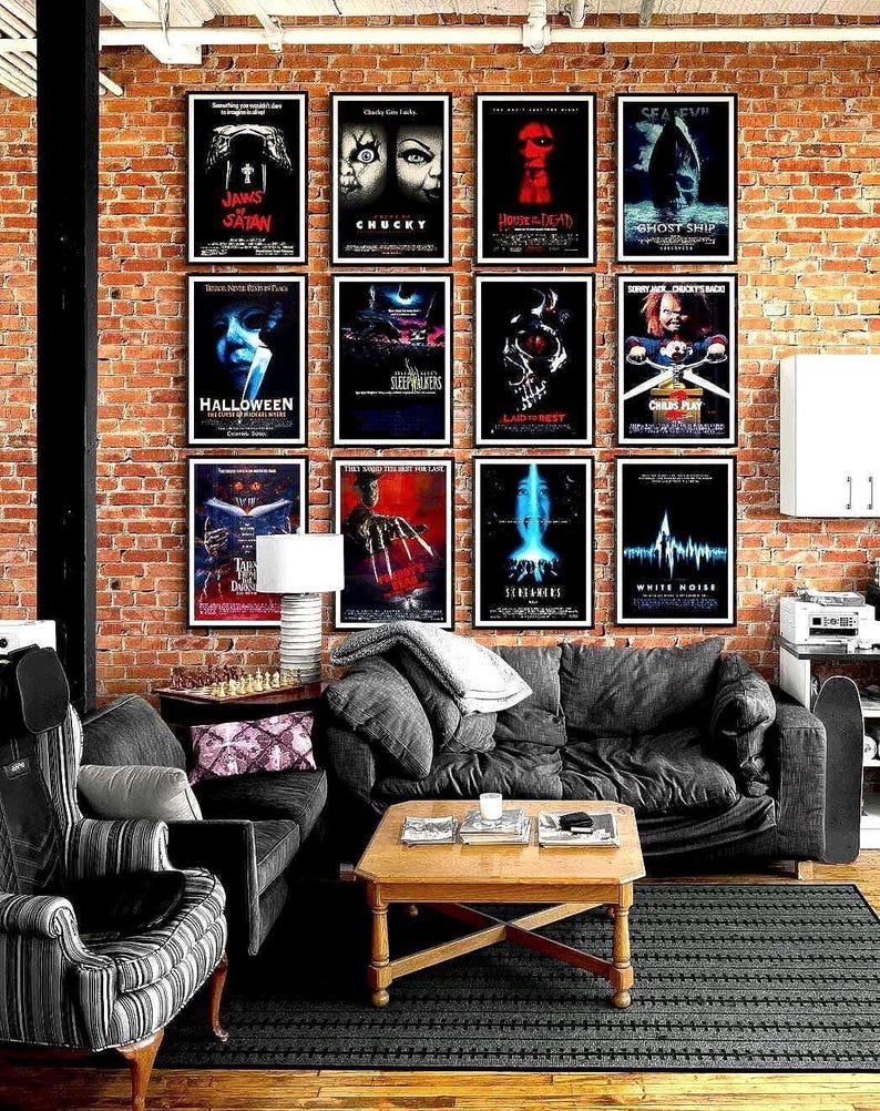 Twelve framed horror movie posters arranged in a grid on a brick wall above a dark grey sectional sofa with blankets, a wooden coffee table and a striped armchair in a cozy living room.