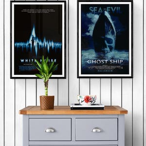 Two movie posters framed in black wood. The poster on the left is for the movie "White Noise" and features a blue and white graphic of a sound wave. The poster on the right is for the movie "Ghost Ship" and features a ship with a skull in the water. The posters are hanging on a white wood wall above a gray dresser.