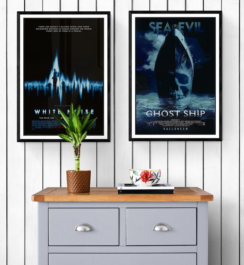Two movie posters framed in black wood. The poster on the left is for the movie "White Noise" and features a blue and white graphic of a sound wave. The poster on the right is for the movie "Ghost Ship" and features a ship with a skull in the water. The posters are hanging on a white wood wall above a gray dresser.