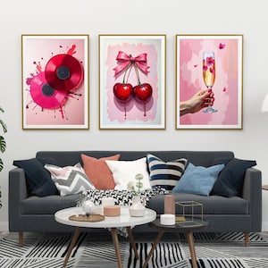 May include: Three framed art prints with pink backgrounds. The first print features two red vinyl records with pink paint splatters. The second print features two red cherries with a pink ribbon. The third print features a hand holding a champagne flute with pink flowers.