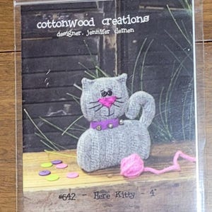 May include: A clear plastic package featuring a gray stuffed cat with a pink nose and a purple collar. The cat is next to a pink ball of yarn and colorful confetti. The text "cottonwood creations" and "Here Kitty - 4" are visible.