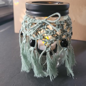 May include: A glass jar with a black lid and a crocheted green cover with black beads. The jar has a small candle inside.