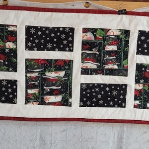 May include: A rectangular quilted table runner with a white background and a red border. The runner features alternating black rectangular panels with white snowflakes and panels with a Christmas-themed pattern. The runner is hanging from clips.