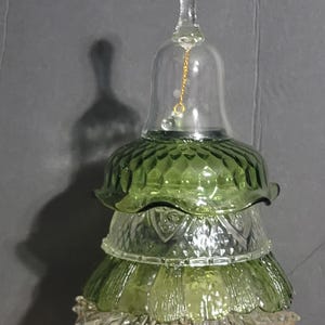 May include: A decorative glass Christmas tree sculpture. The tree is constructed from stacked glass bowls in shades of green and clear, topped with a bell-shaped glass finial. A gold chain hangs inside the bell. The tree sits on a clear glass base.